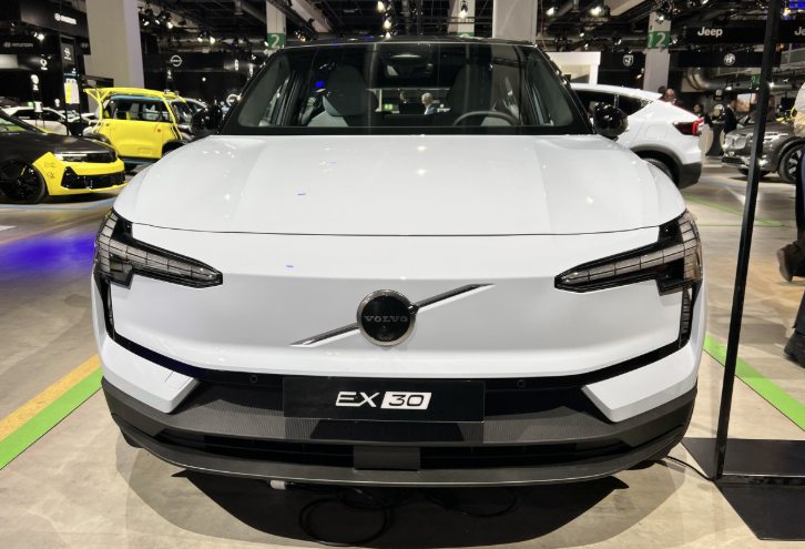 front view of a white Volvo EX30 electric car displayed indoors at an auto show with other vehicles in the background