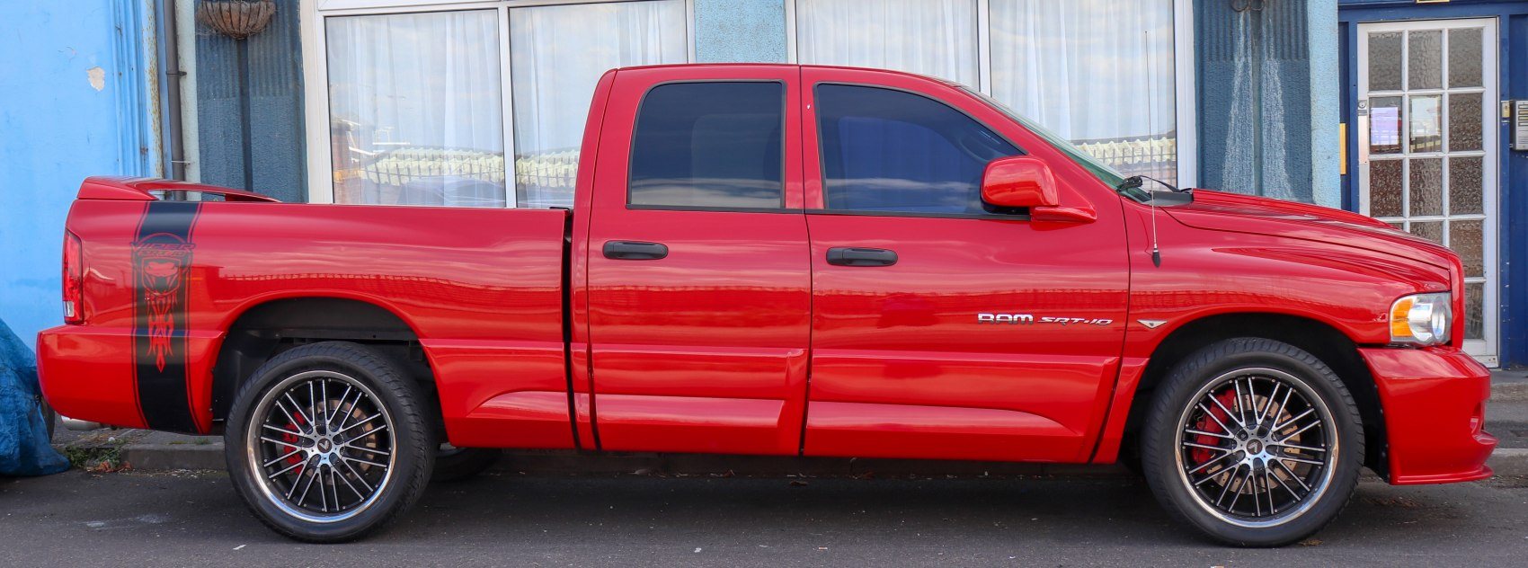 side view of a red Dodge Ram SRT-10 pickup truck with black racing stripes and custom wheels parked in front of a blue building with large windows and a glass door