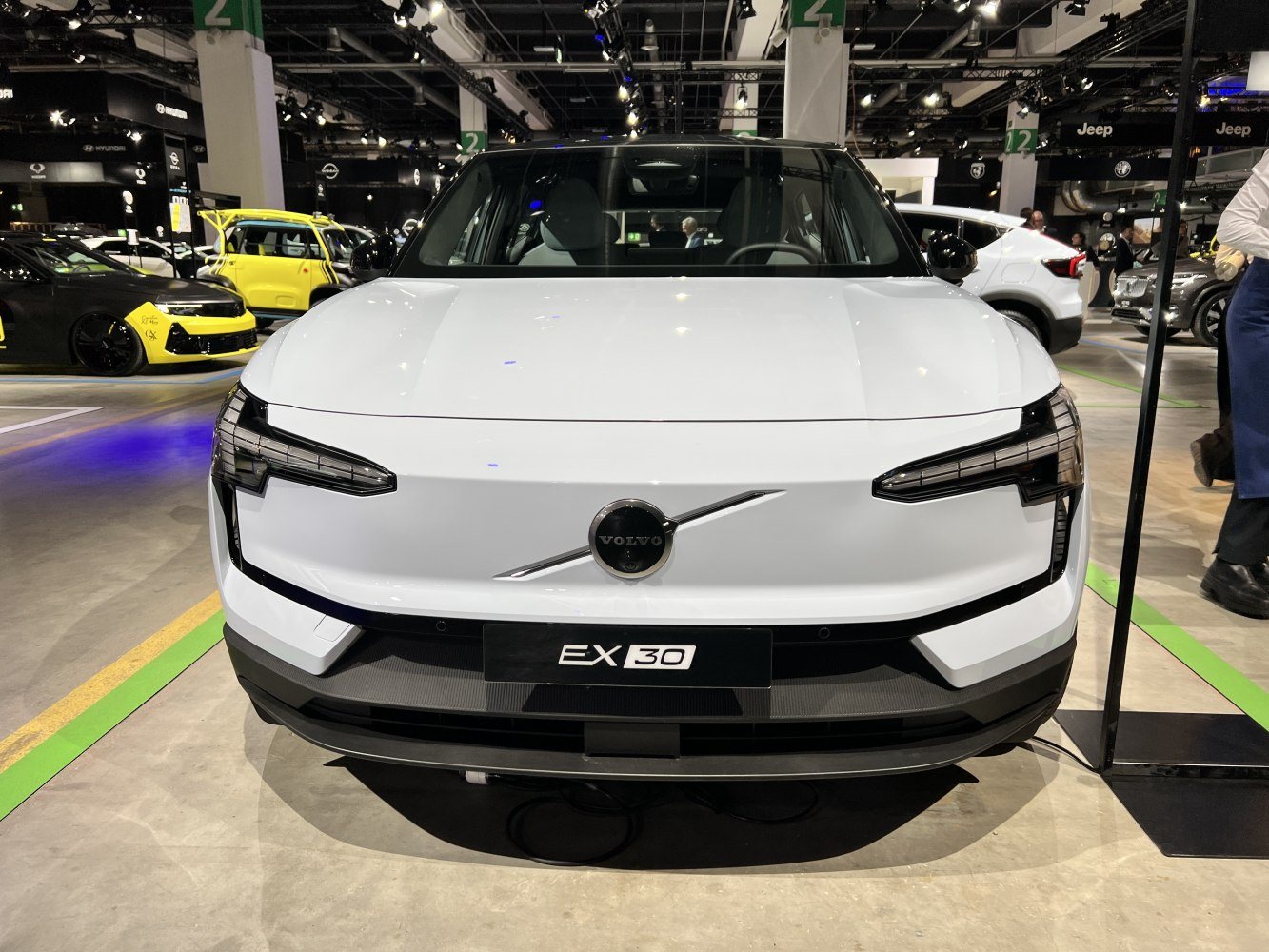 front view of a white Volvo EX30 electric car displayed indoors at an auto show with other cars and people in the background
