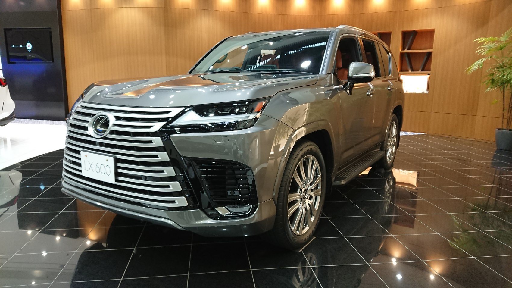 front side view of a grey Lexus LX 600 luxury SUV displayed indoors on a black tiled floor with wooden wall background