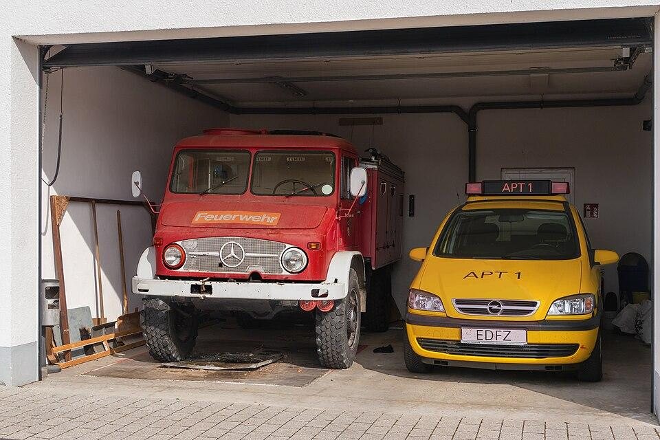 Red vintage Mercedes fire truck and yellow Opel car parked side by side in a garage