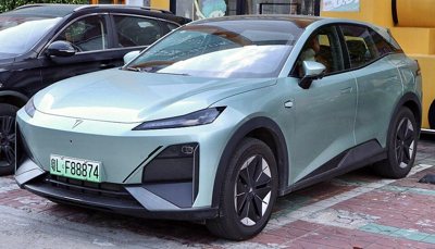 Front side view of a light green modern electric SUV parked on a brick-paved street with distinctive angular headlights and black wheel rims