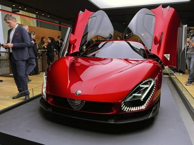 front view of a shiny red sports car with open butterfly doors displayed at an indoor car show with people in the background