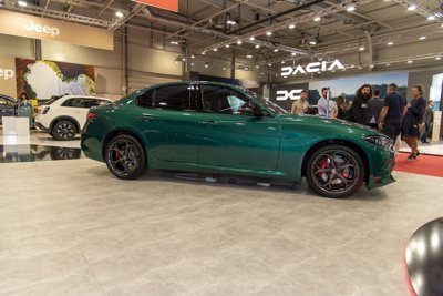 Green luxury sedan displayed indoors at a car show with Jeep and Dacia booths in the background and people walking around