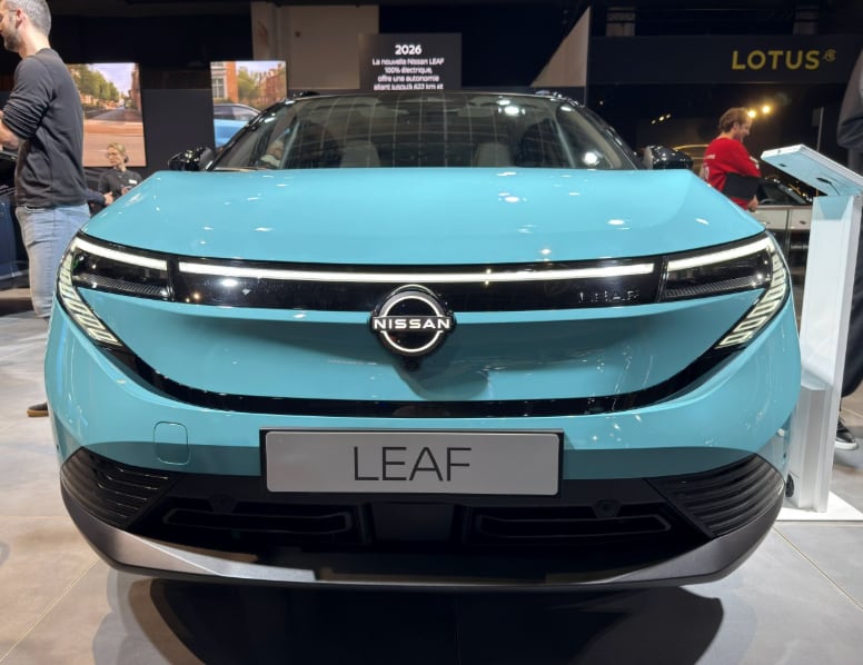 front view of a light blue 2026 Nissan Leaf electric car displayed at an auto show with visible Nissan logo and Leaf nameplate