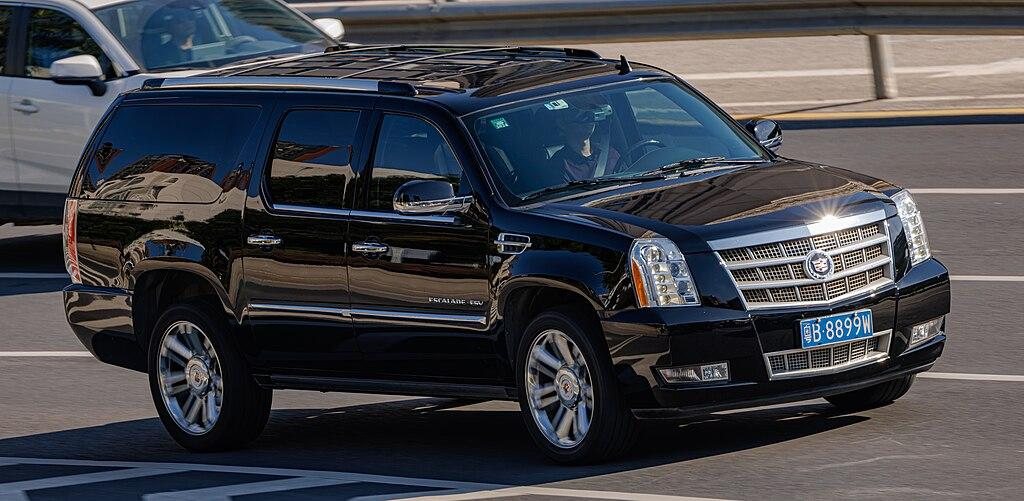 Black Cadillac Escalade SUV driving on a multi-lane road during daytime with a white vehicle in the background