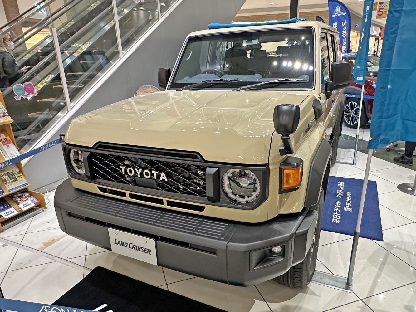 front view of a beige Toyota Land Cruiser SUV displayed indoors at a showroom with tiled floor and escalator in the background