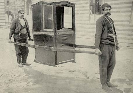 black and white image of two men carrying a traditional enclosed palanquin sedan chair with wooden poles