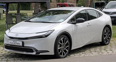 front side view of a white Toyota Prius plug-in hybrid car parked on a cobblestone driveway with green grass and trees in the background