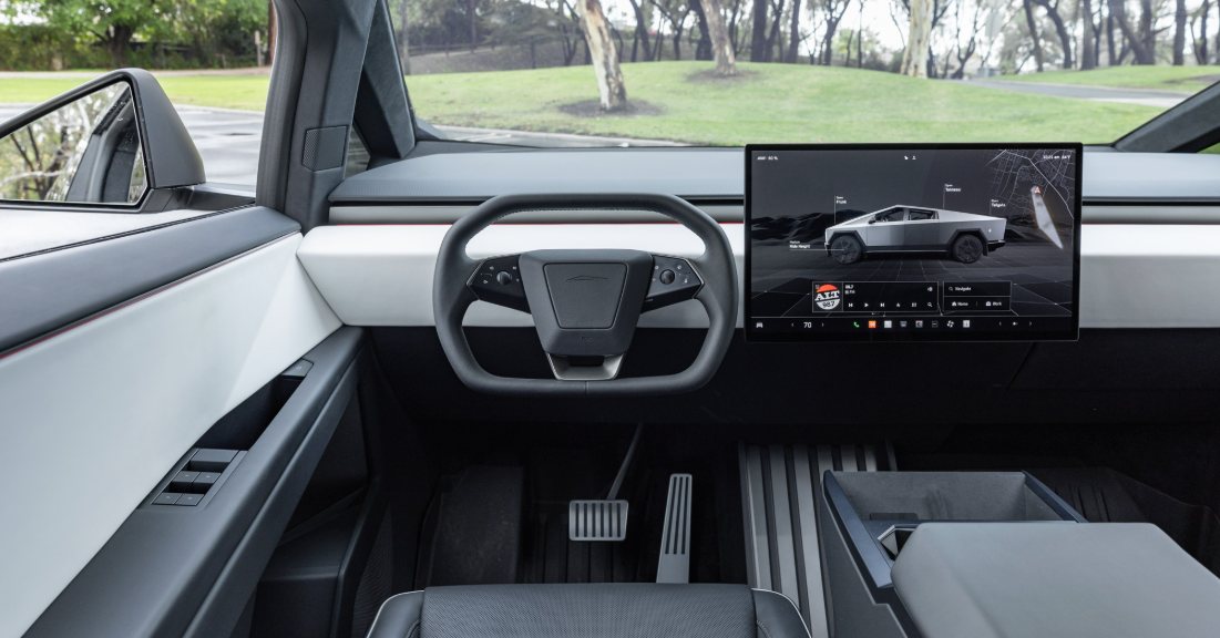 Interior view of a modern electric vehicle with a minimalist dashboard featuring a digital steering wheel and a large central touchscreen display showing a 3D model of a futuristic truck