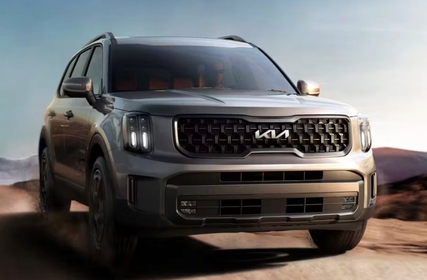 front view of a gray Kia SUV driving on a dirt road with mountains in the background