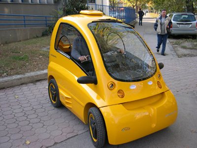 Small yellow compact electric car parked on a sidewalk with a person inside and two pedestrians walking nearby