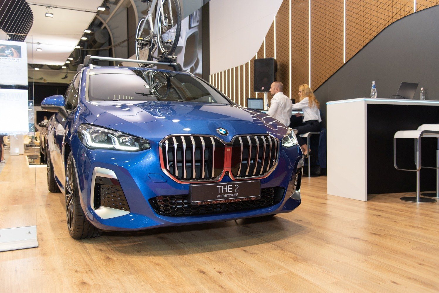 front view of a blue BMW THE 2 Active Tourer displayed indoors with a bicycle mounted on the roof rack and two people sitting in the background