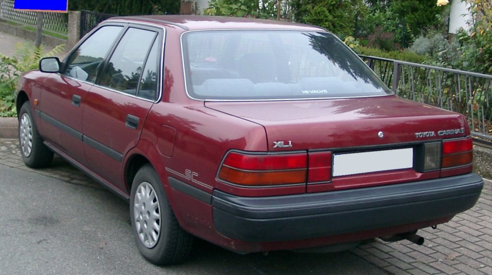 Rear and side view of a red sedan with rectangular tail lights and black bumper