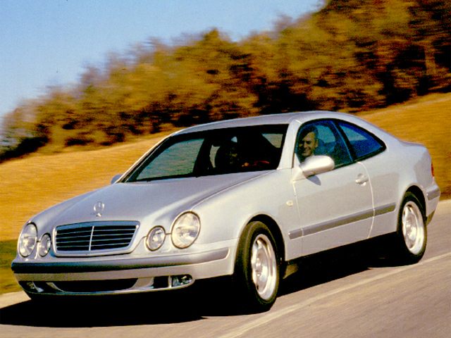 Silver classic coupe with dual round headlights driving on a rural road.