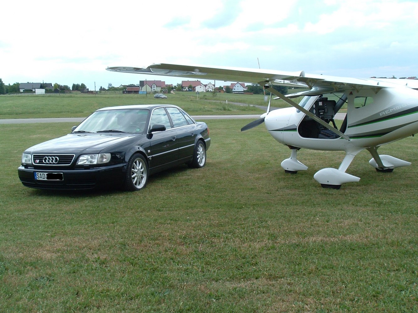 Black sedan parked next to light aircraft on grassy field