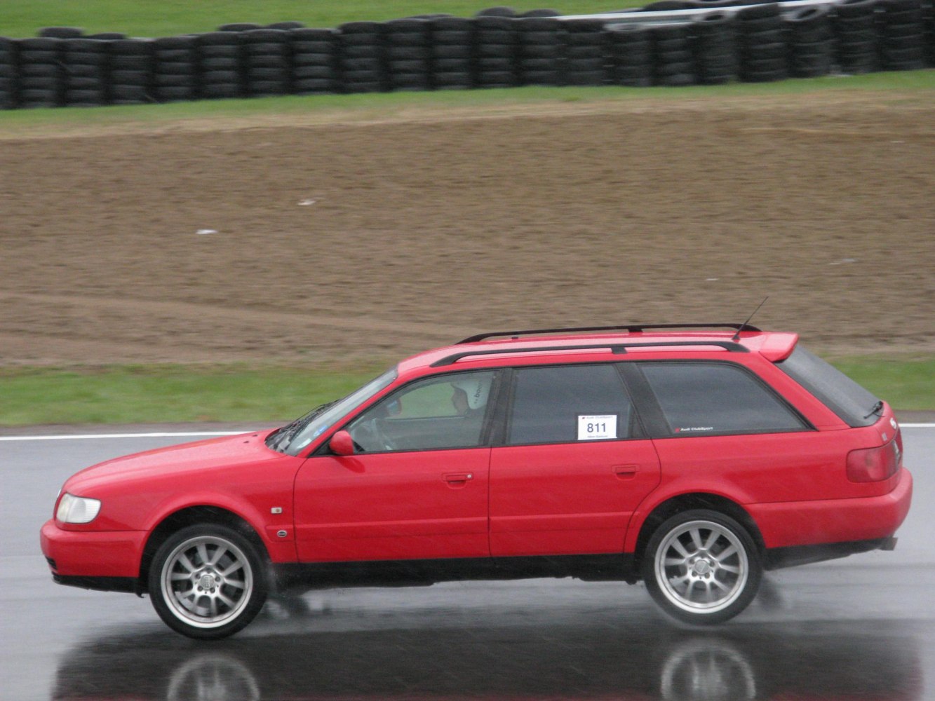 Red station wagon with roof rails driving on wet racetrack.