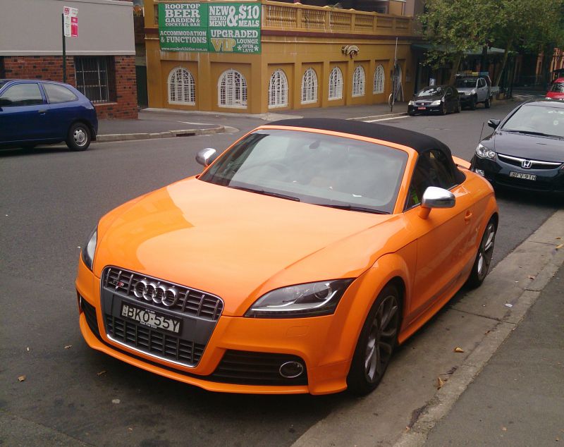 Orange convertible with black soft top and silver mirrors from front side.