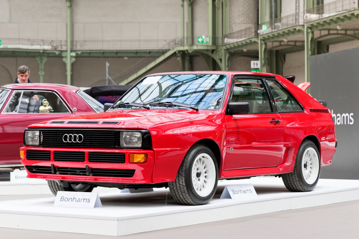 Red classic rally car with boxy design, front grille, and white multi-spoke wheels