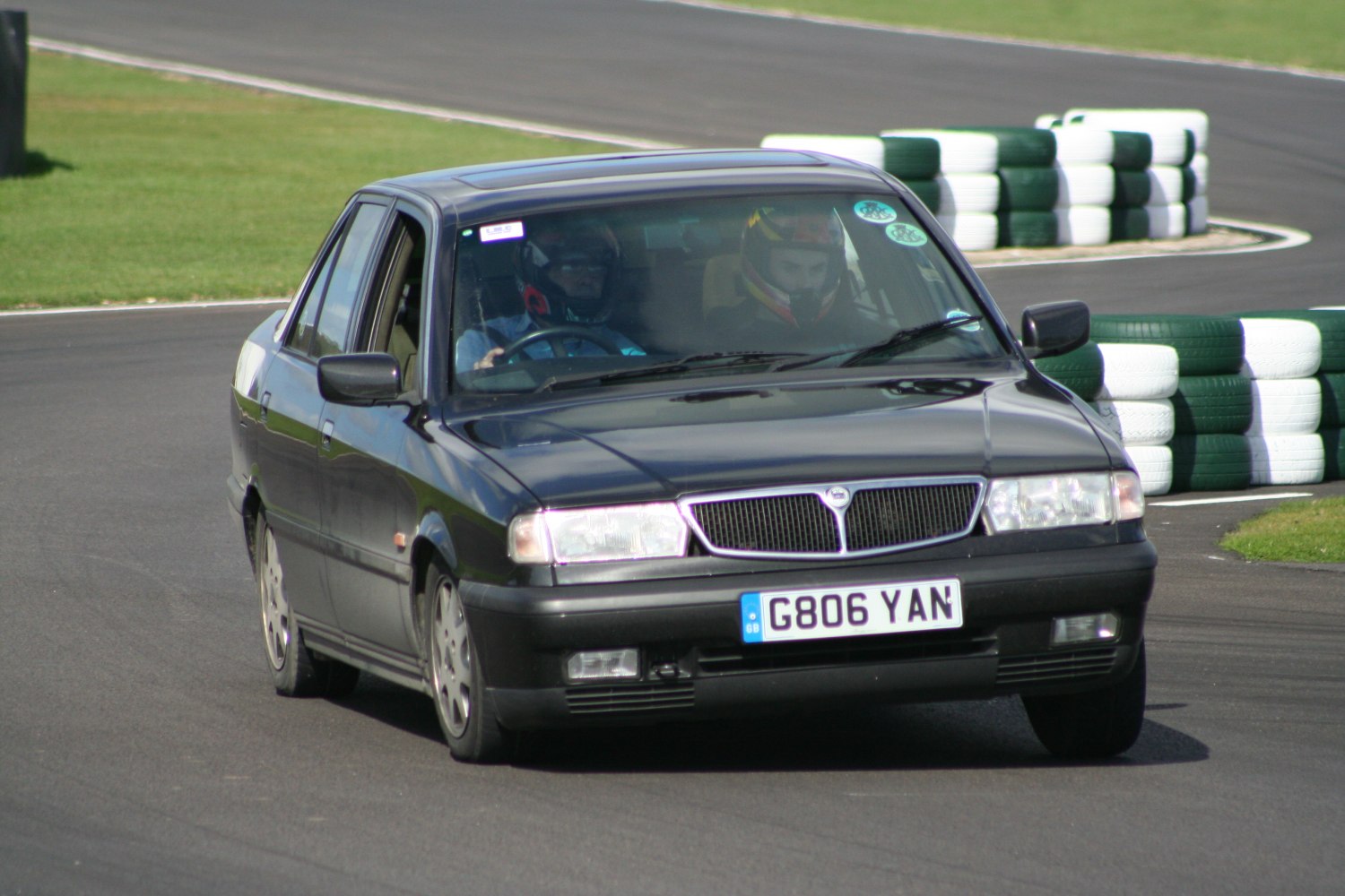 Classic 1990s saloon front view with helmeted driver and passenger on racetrack.