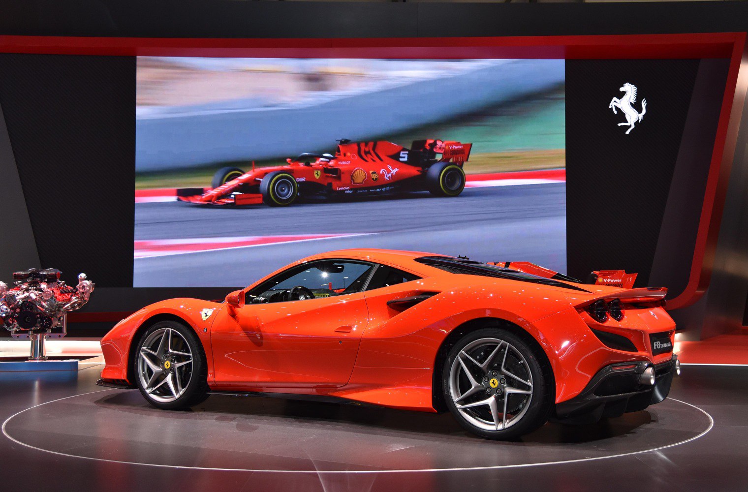 Side profile of bright orange sports car with visible engine and racing video background