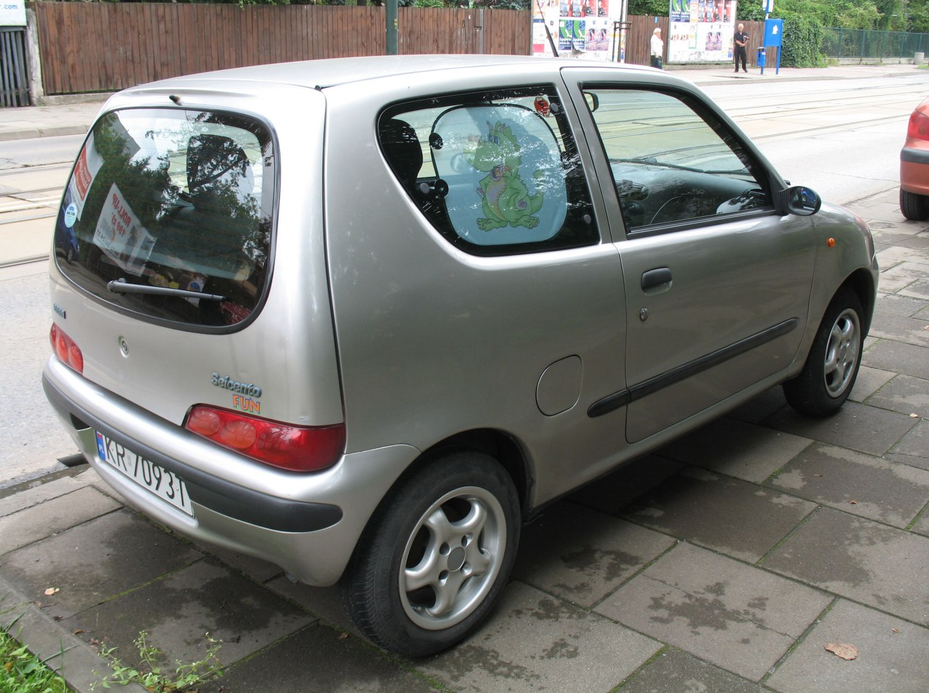 Rear and side of a silver compact hatchback with a cartoon sunshade on a small rear side window.