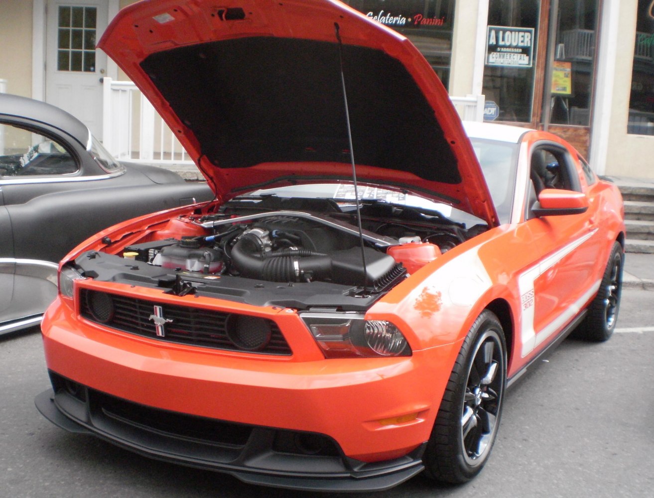 Front view of a Mustang V with open hood showing engine and black wheels