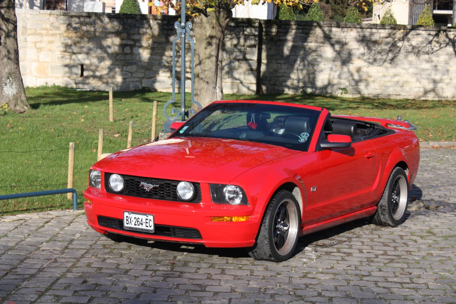 Red convertible with front grille and chrome wheels on cobblestone.