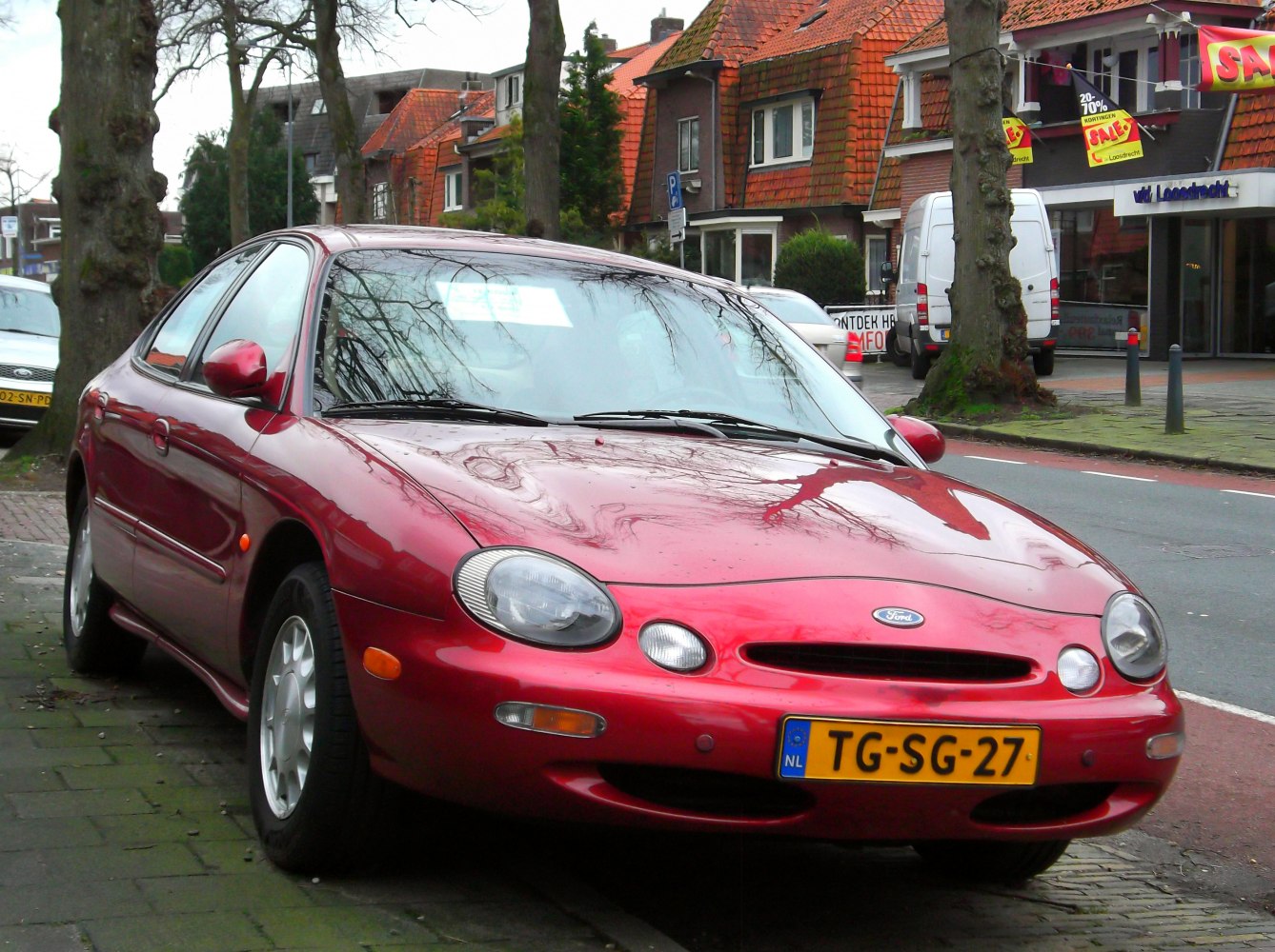 Red sedan with oval headlights and aerodynamic body parked on street curb