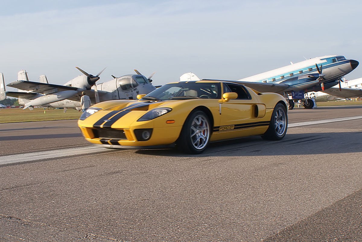 Yellow supercar with black racing stripes on airport runway near vintage planes.
