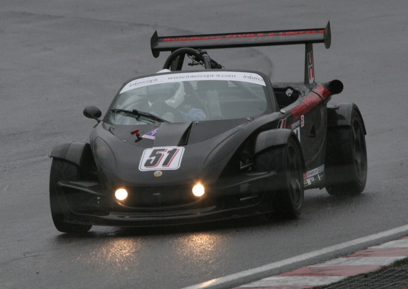 Open-top sports car with large rear wing and headlights active on wet racing track