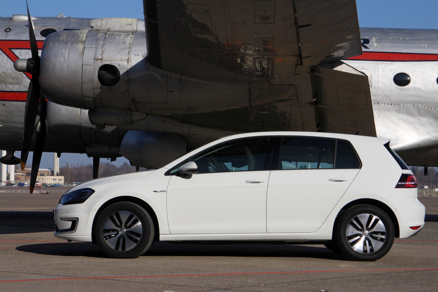 White 5-door hatchback with alloy wheels, vintage propeller airplane in background