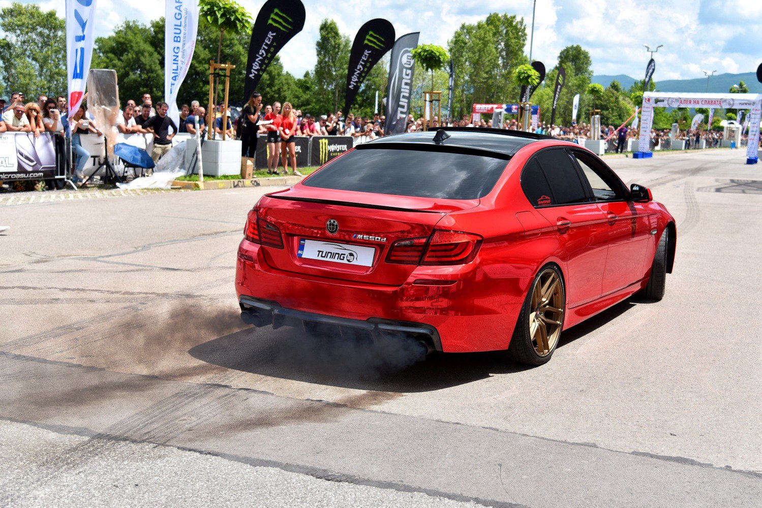 Red sedan with black tinted windows and gold wheels emitting burnout smoke.