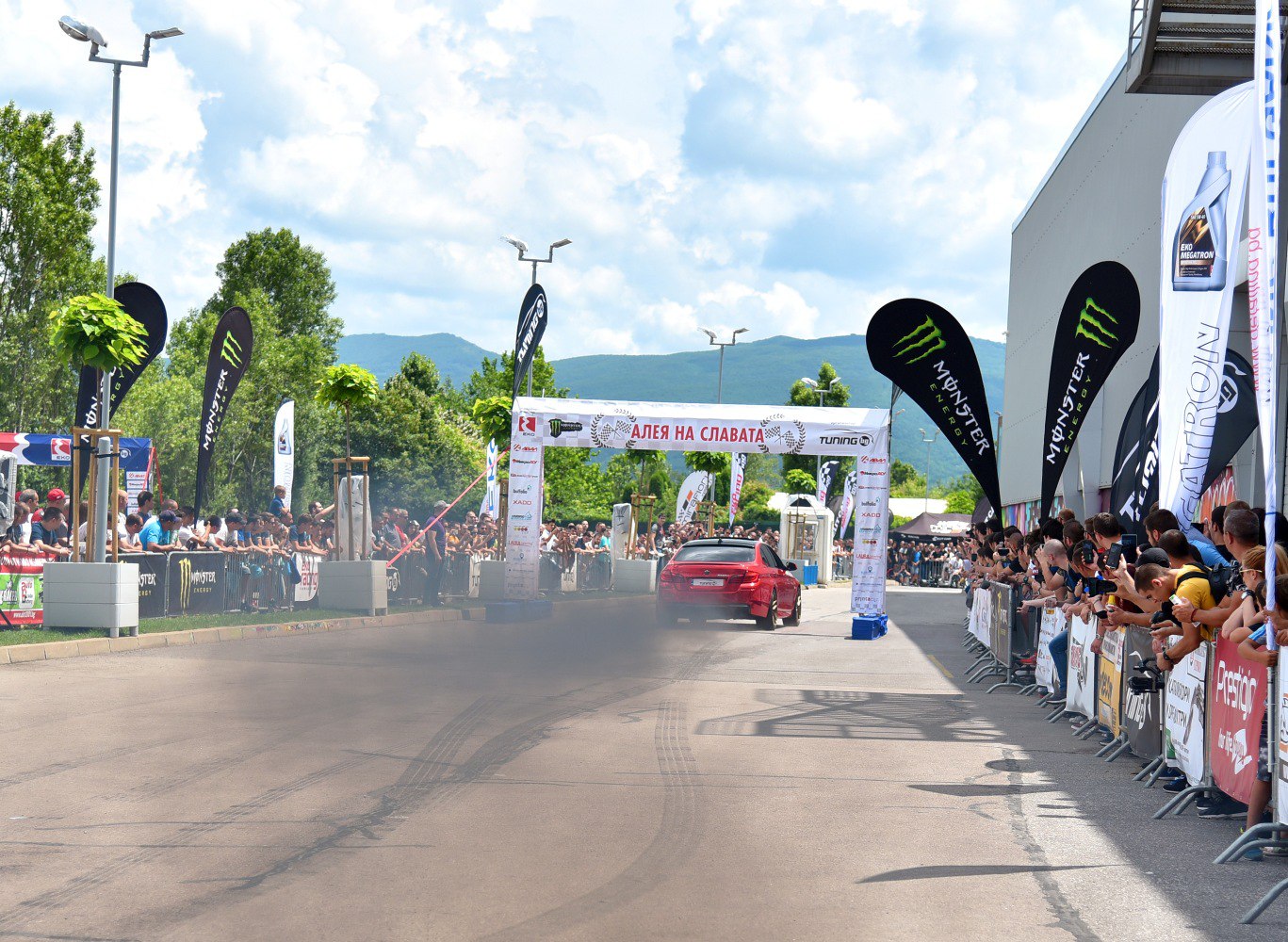 Red sedan performing a tire burnout with smoke amid crowd at motorsport event