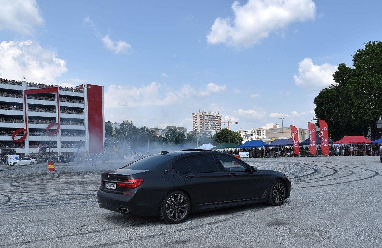 Matte black sedan drifting on an open track with tire smoke and crowd.
