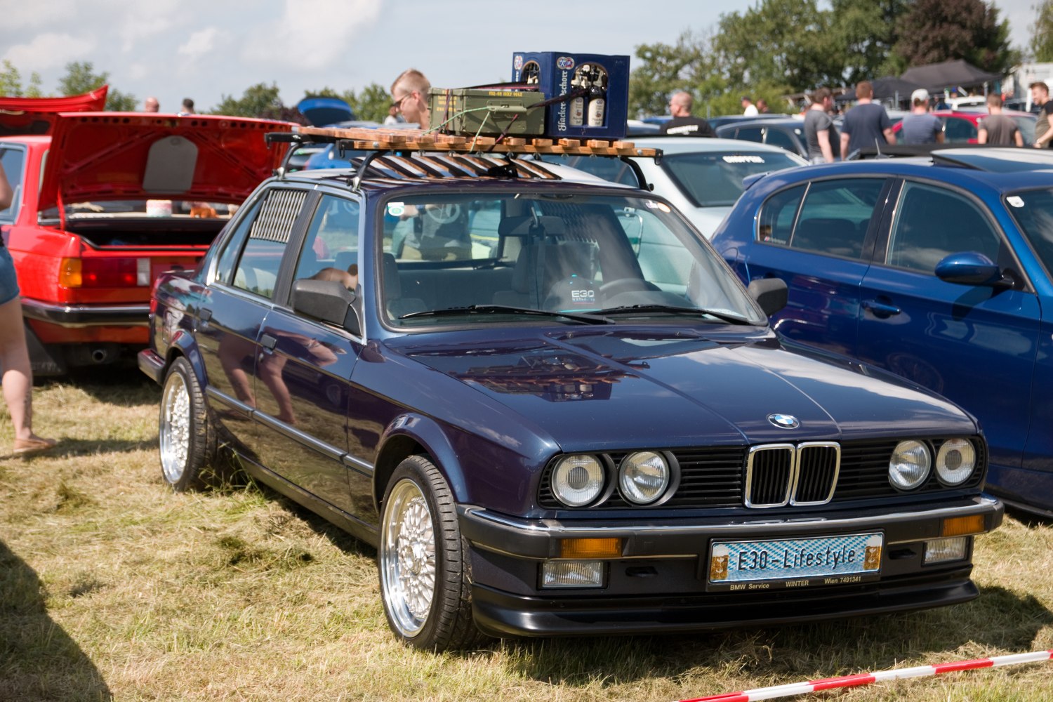 Classic four-door sedan with round headlights, polished multi-spoke wheels, and roof rack loaded with wooden pallet and bottles.