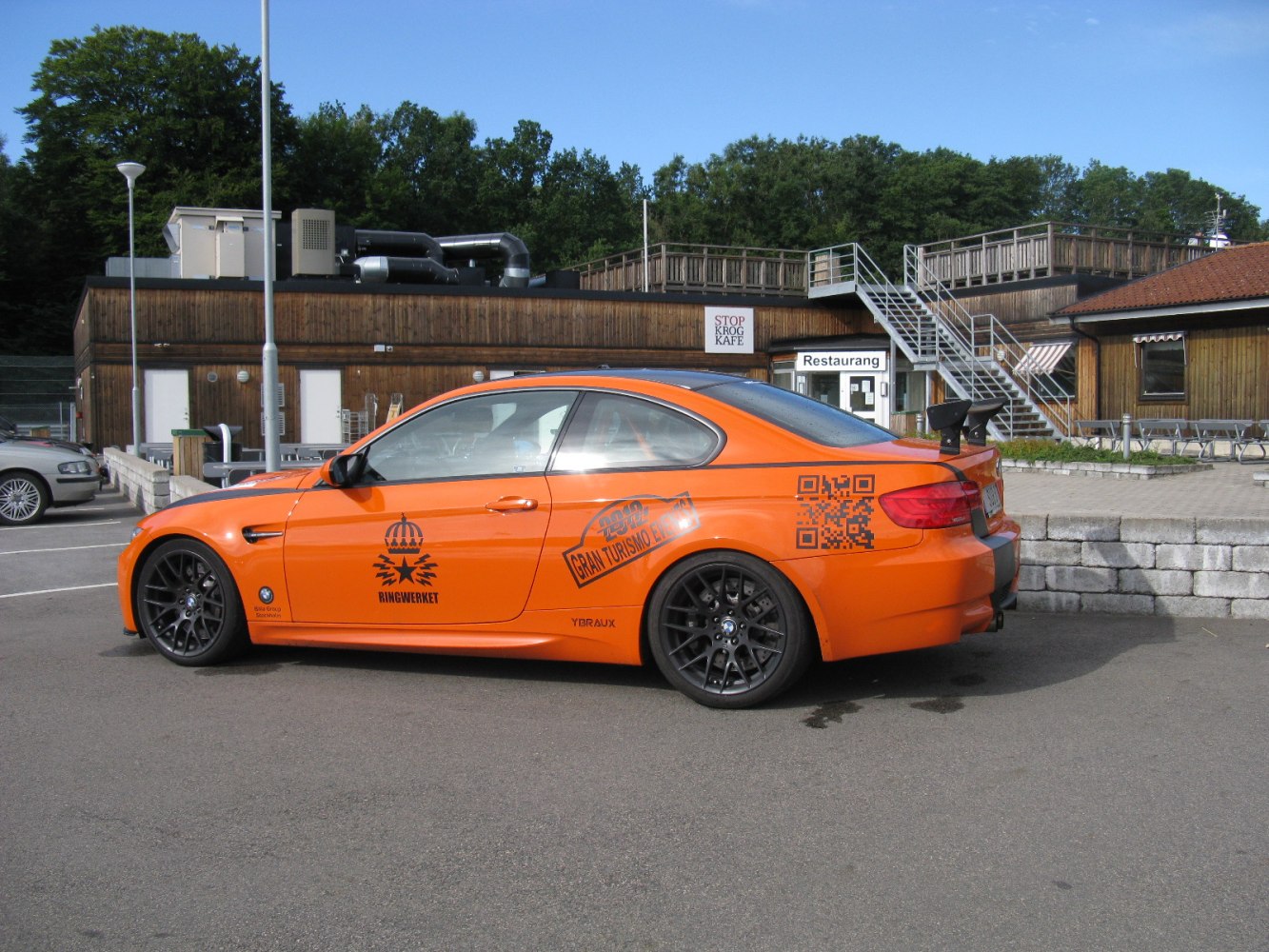 BMW M3 coupe with orange body, racing decals, black wheels, and rear wing spoiler