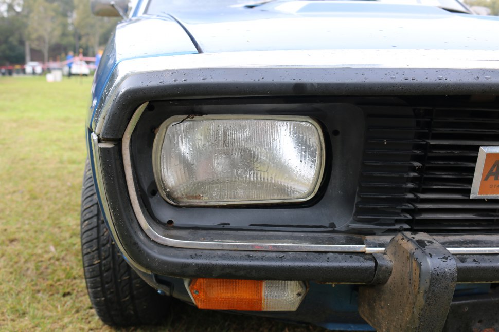 Close-up of vintage car's rectangular headlight, grille, and bumper with worn details.