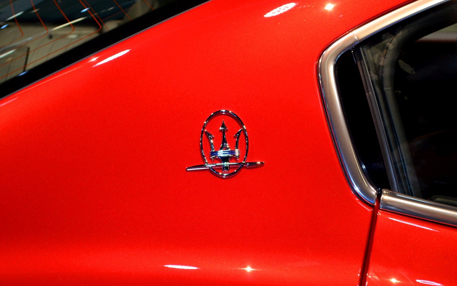 Close-up of Maserati trident emblem on red side panel with chrome window trim