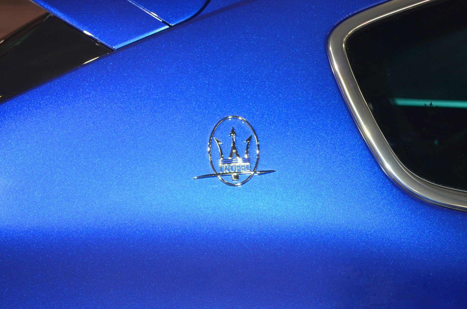 Close-up of a silver Trofeo side emblem on metallic blue paint near a window.