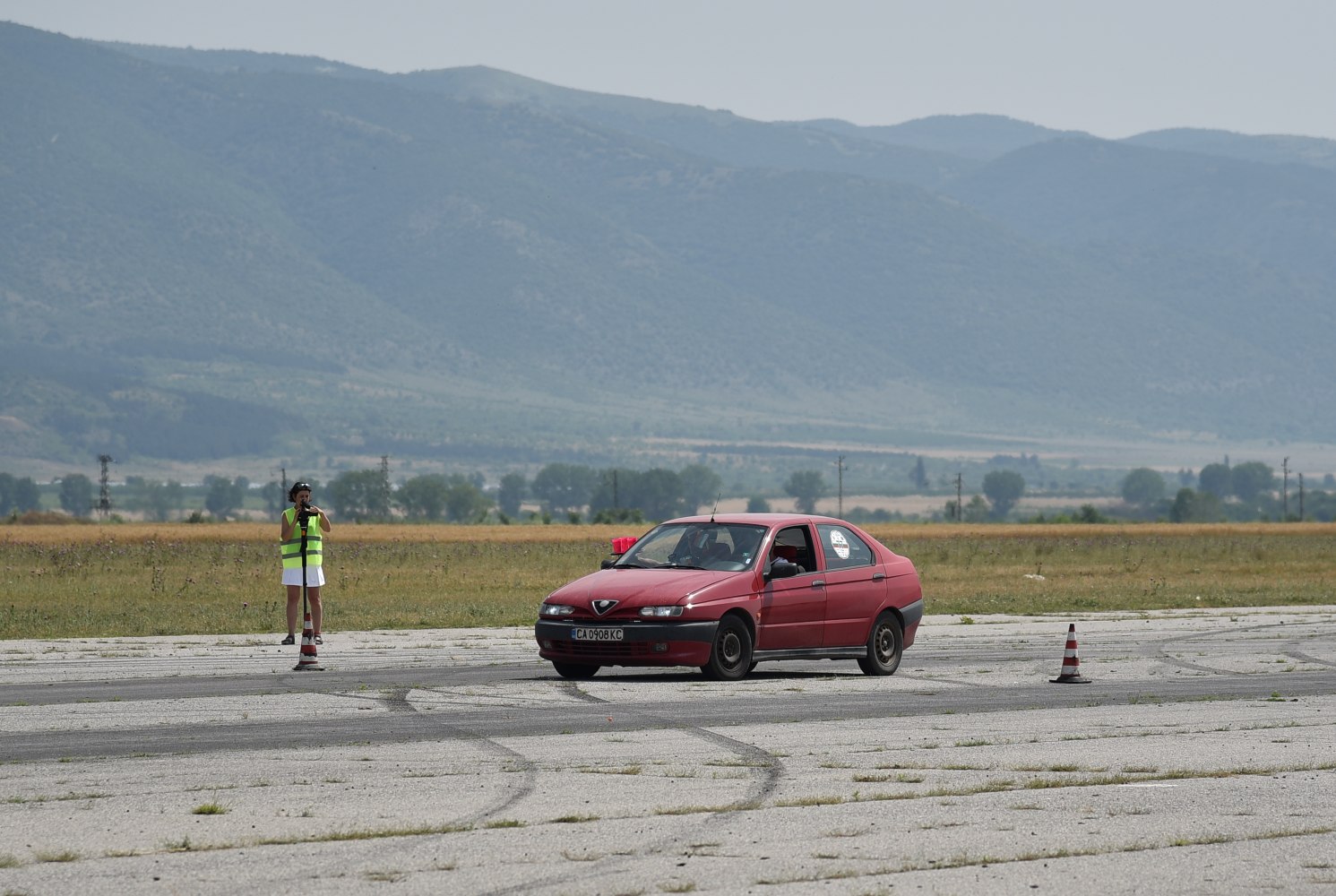 Red sedan navigating traffic cones on cracked asphalt with mountain backdrop