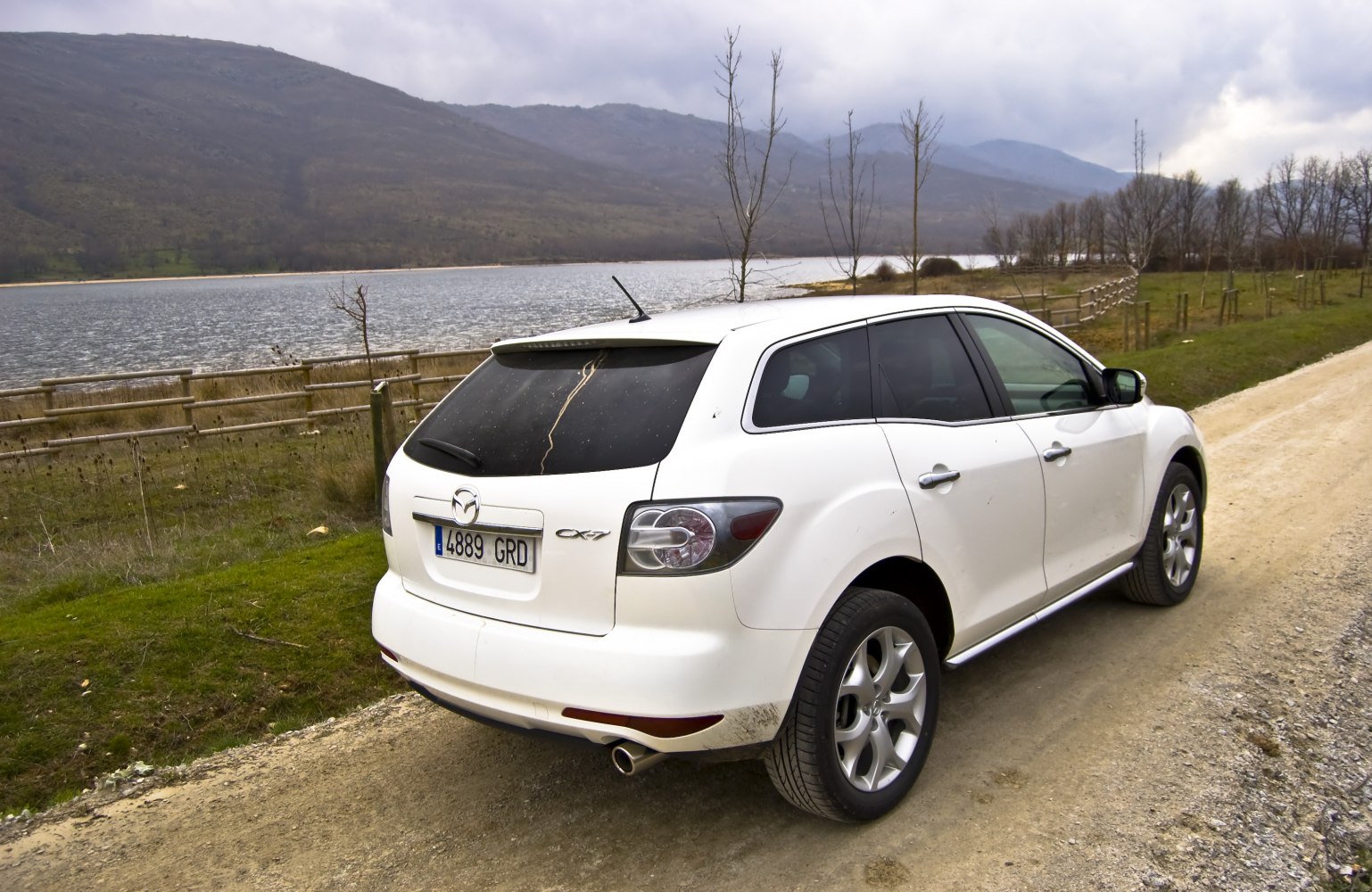 Rear view of white SUV on dirt road by lake and hills