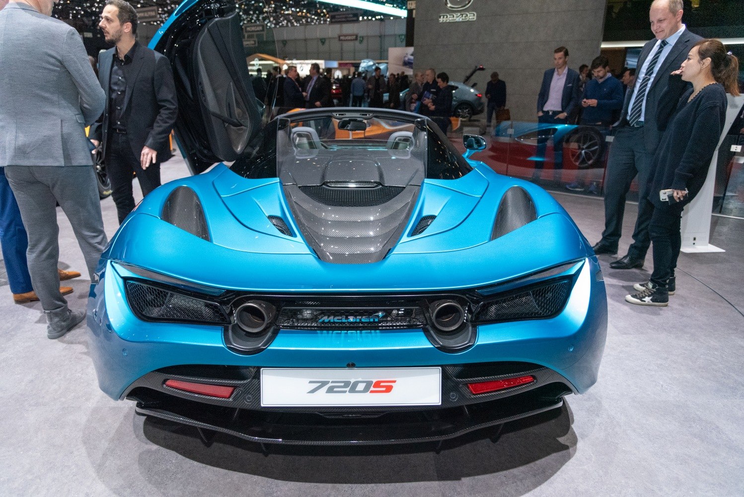 Rear view of a blue convertible sports car with open upward-opening doors and exposed carbon fiber engine cover