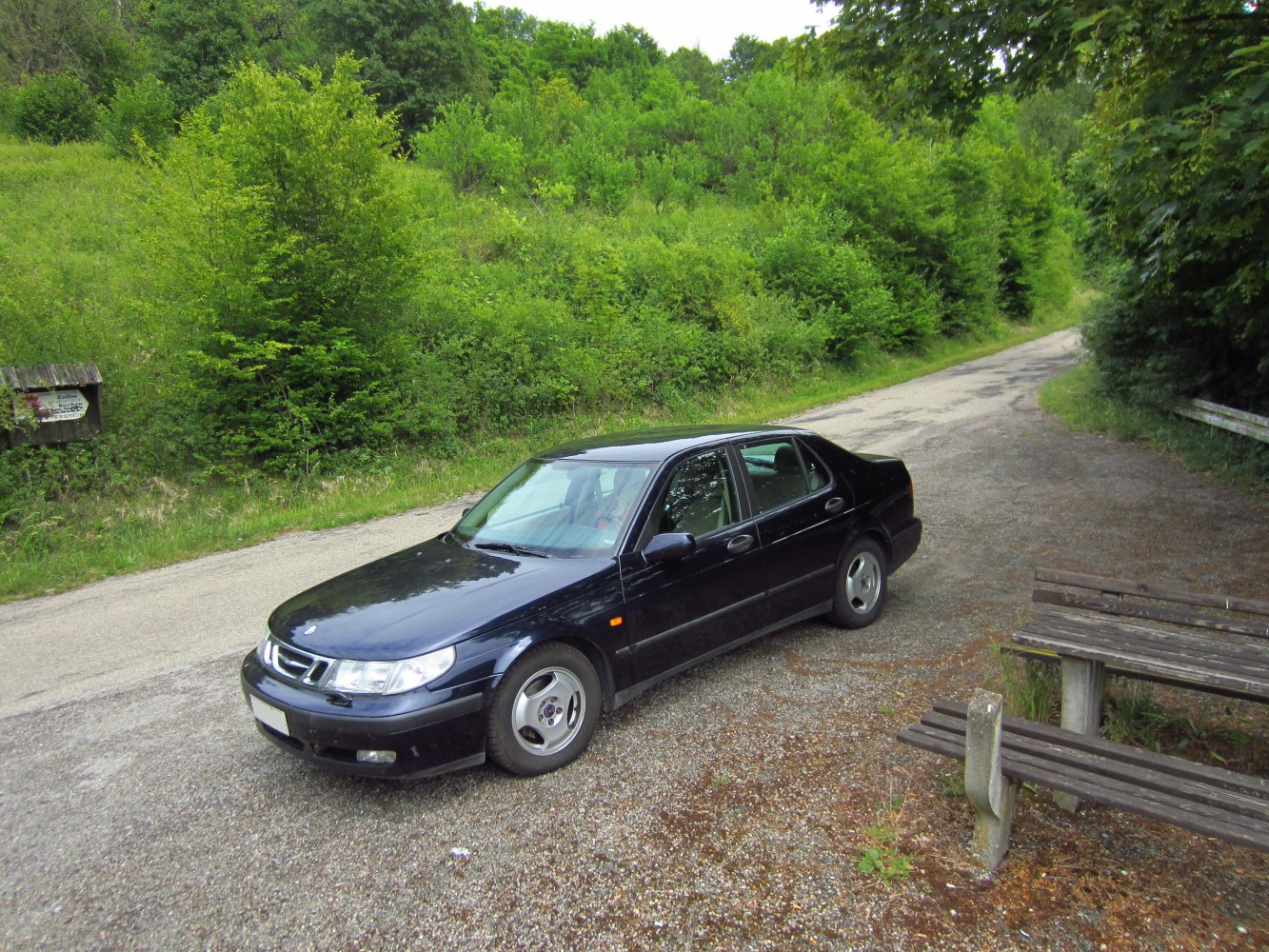 Black four-door sedan parked near a forest road with wooden benches nearby