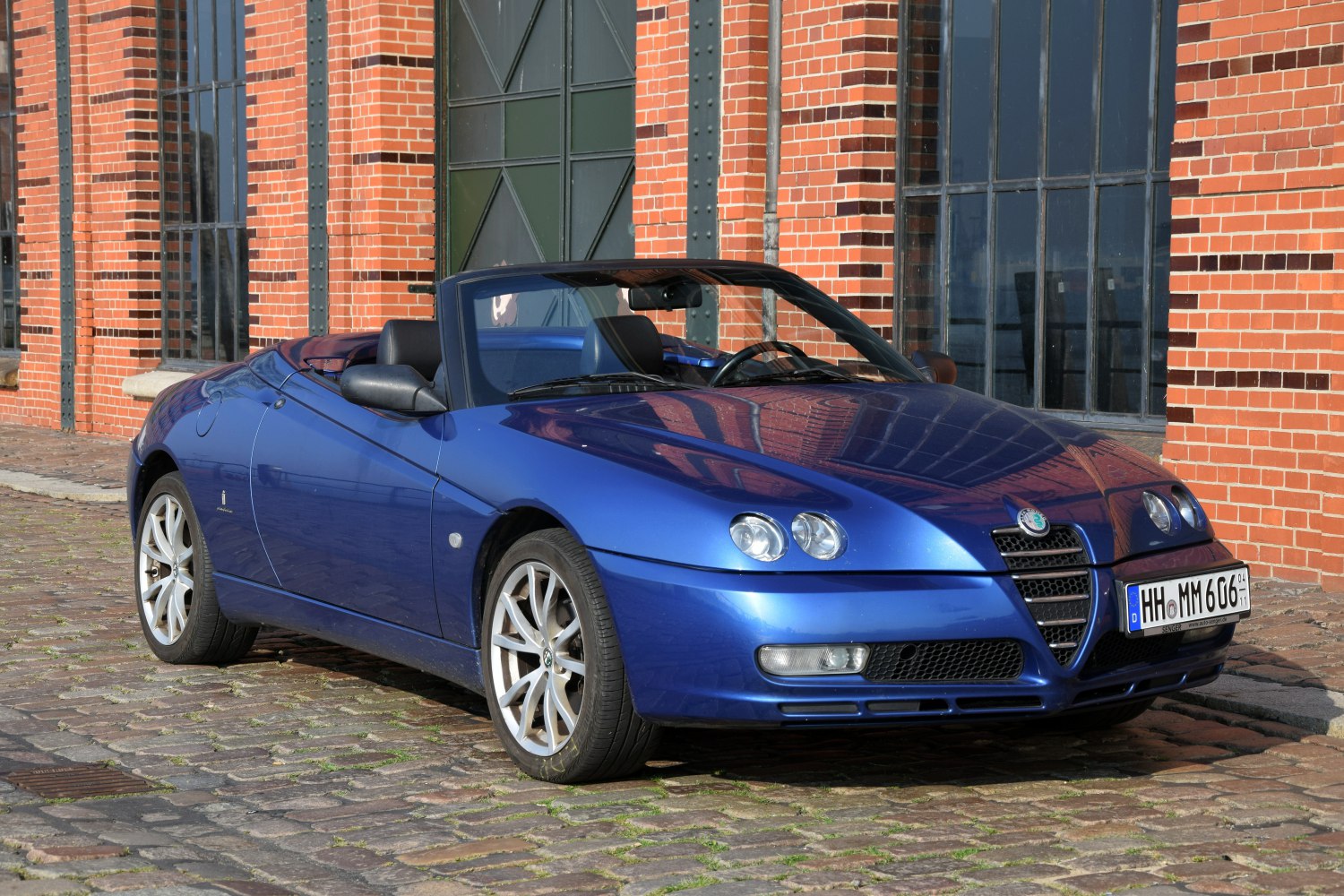 Blue convertible with dual round headlights and alloy wheels on cobblestone.