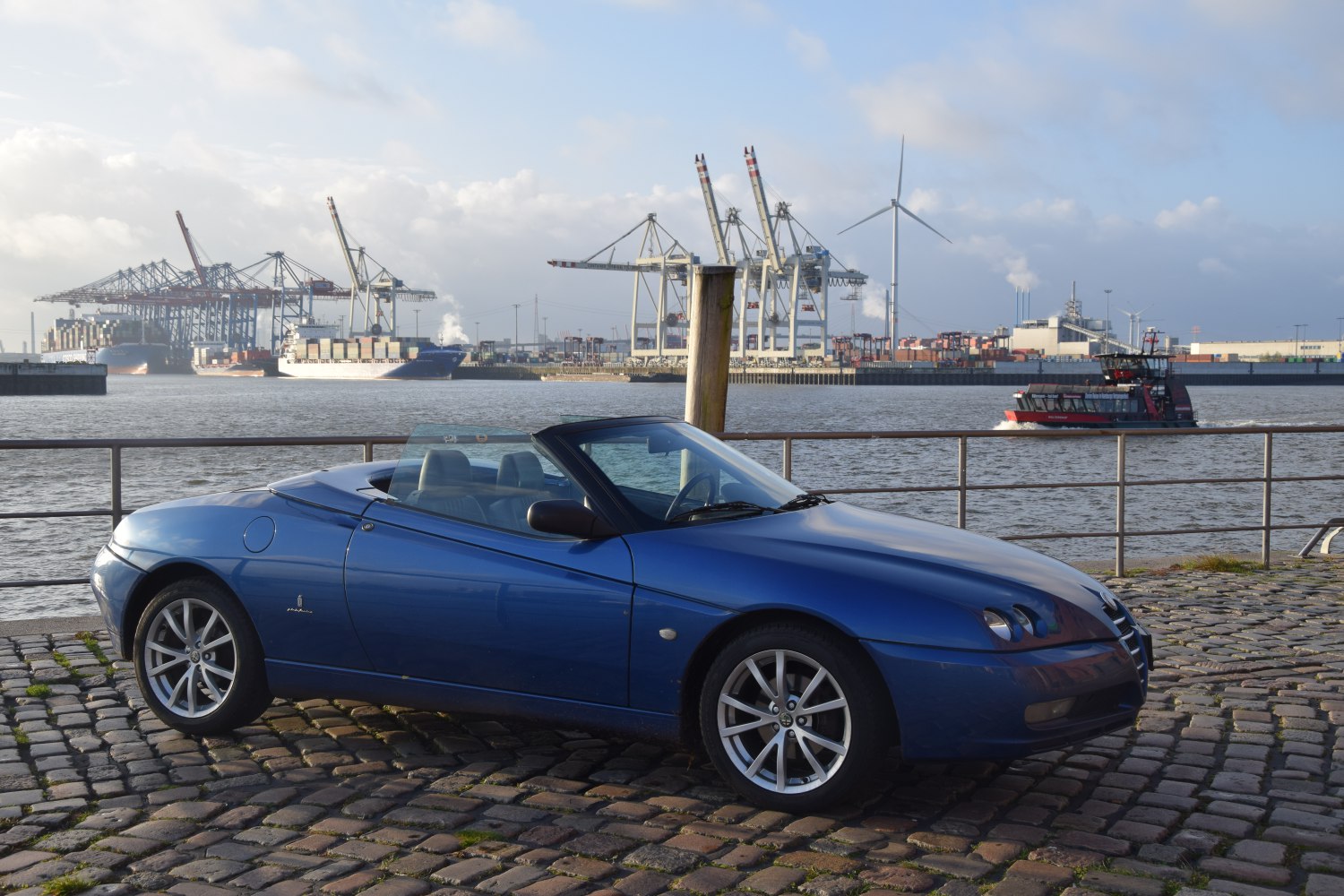 Blue convertible sports car with soft top down parked by waterfront.