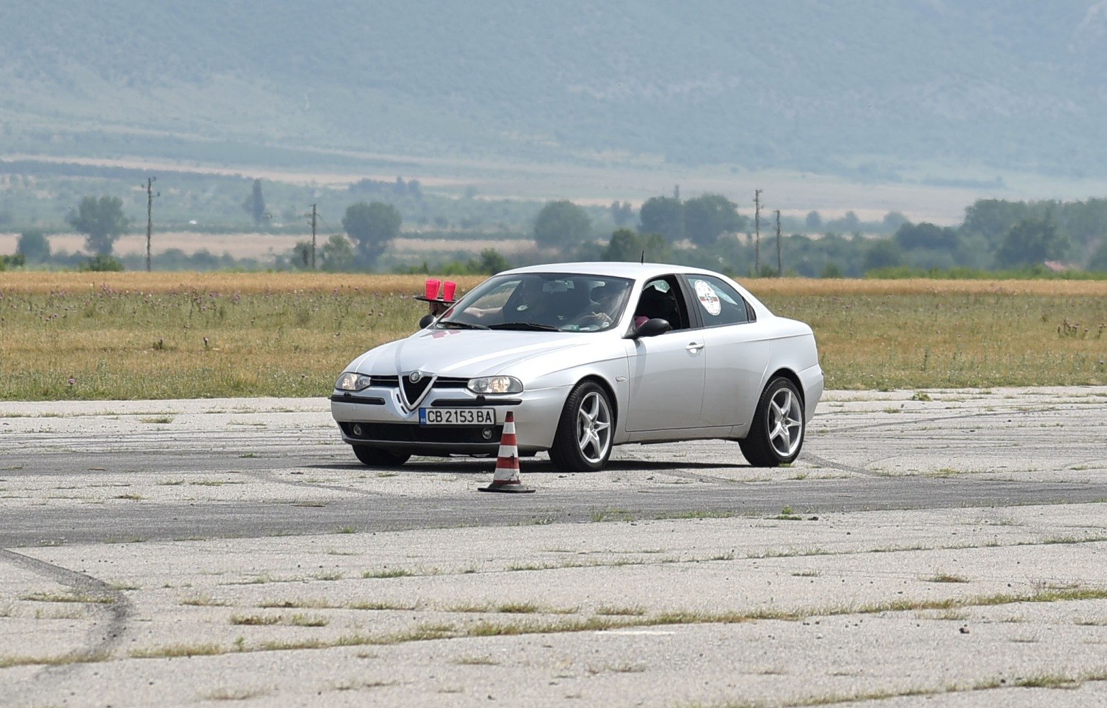 Silver sports coupe with dual round headlights and V-shaped grille on paved open area.