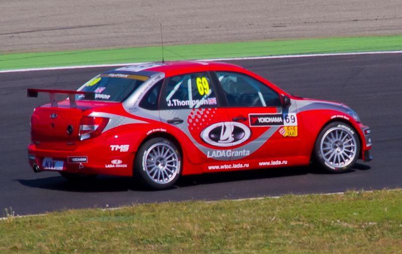 Red racing sedan with decals and rear spoiler on racetrack.