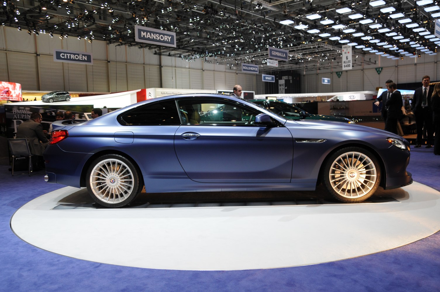 Side profile of matte blue luxury sport coupe with multi-spoke alloy wheels on display platform.