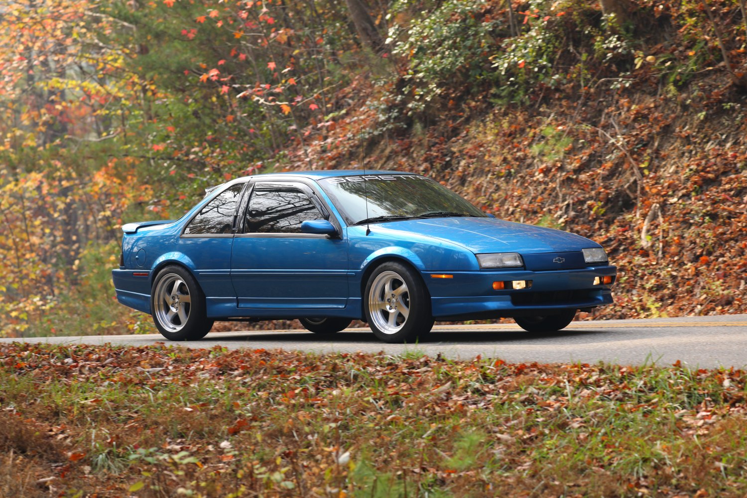 Blue sports coupe side-front view on forest road with fall leaves.